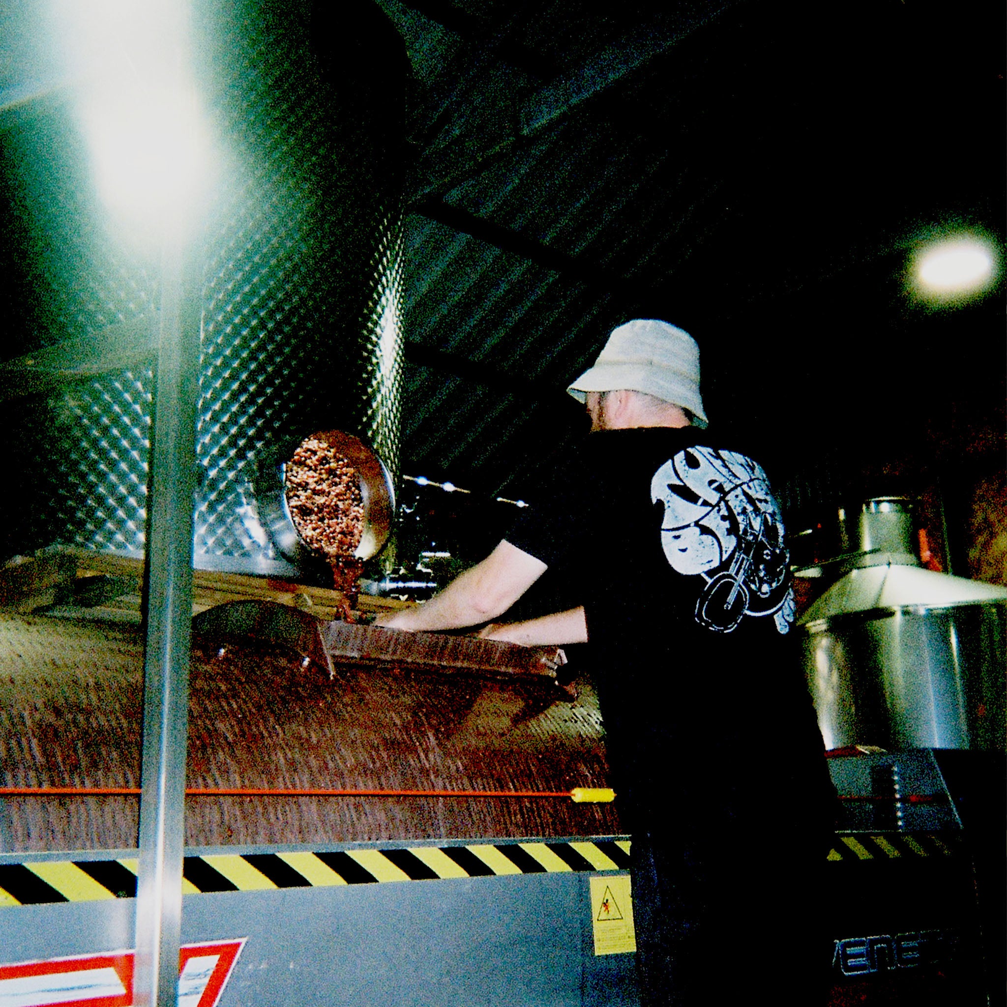 Person in a black t-shirt and white bucket hat standing in front a stainless-steel grape press inside a winery.