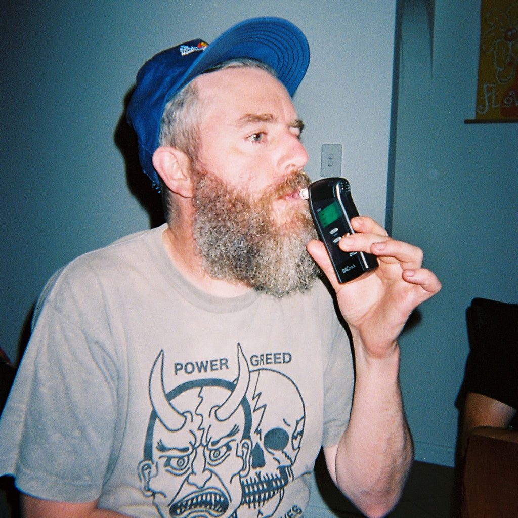A person with a beard, wearing a beige t-shirt and a blue cap, blowing into a personal breathalyser machine.