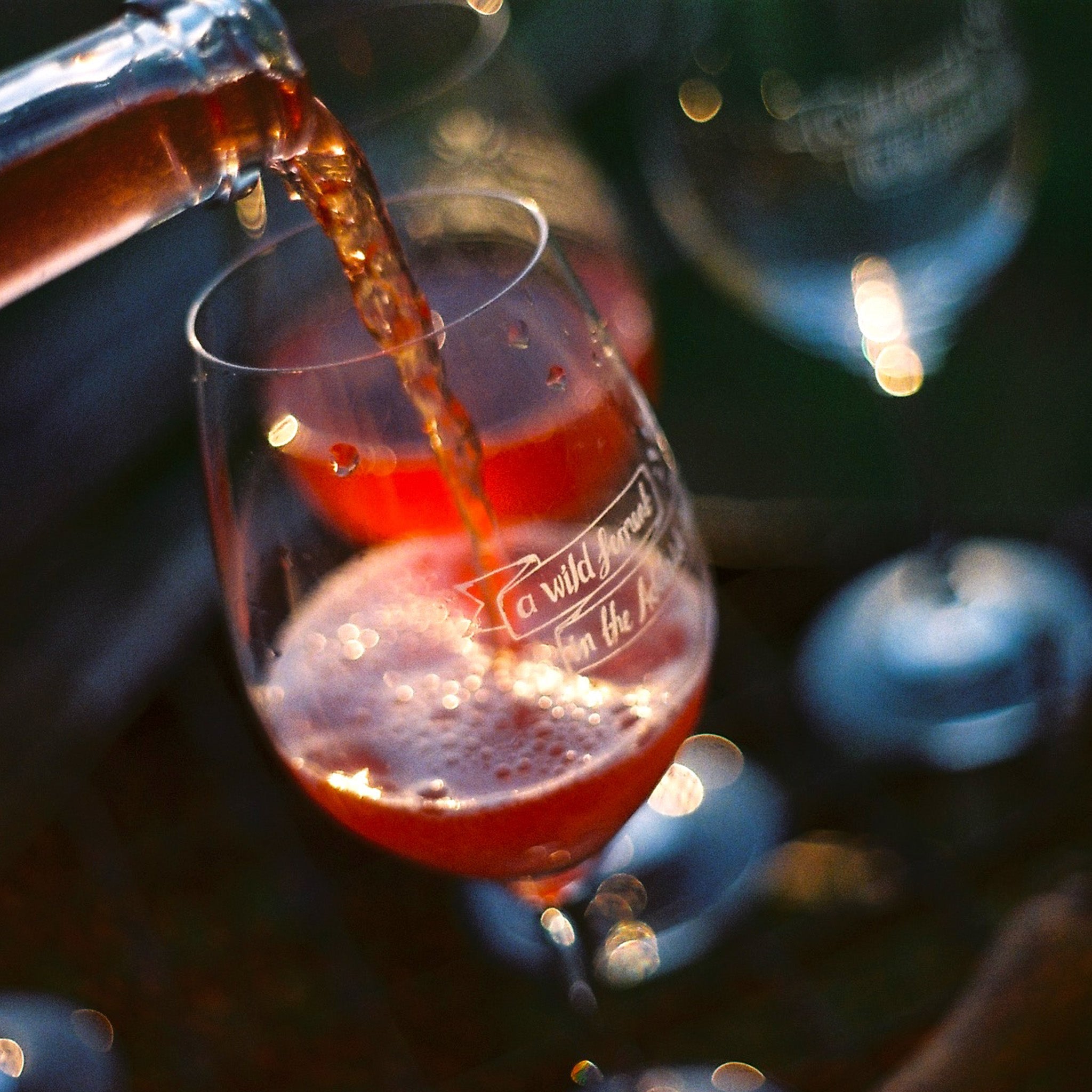 A close up of Borachio Pash Rash 2024 being poured from a bottle into a wine glass, with empty glasses softly visible in the background.